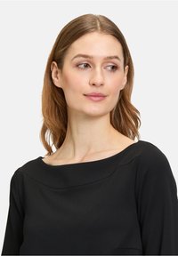 Schwarze Bluse mit rundem Halsausschnitt, langen Ärmeln und glattem Stoff. Das Design hat dezente Nähte im Brustbereich.