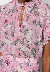 Blusa floral rosa con acentos en negro y amarillo, que presenta un escote en forma de llave y lazos, mangas cortas y una tela ligera y texturizada.
