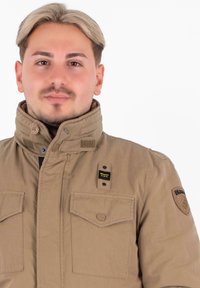 Giovane uomo con capelli castano chiaro che indossa una giacca Blauer beige con colletto alto e numerose tasche con bottoni, rivolto in avanti.