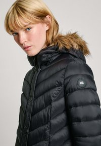 Zwarte pufferjack met een ritssluiting, horizontale stiksels en een capuchon met nepbontaccenten. Logopatch op de linker mouw.