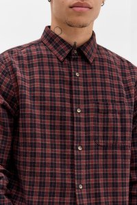 Homem a usar uma camisa de botão xadrez vermelha e preta com bolso no peito, tatuagens visíveis no pescoço e um pequeno brinco em argola na orelha esquerda.