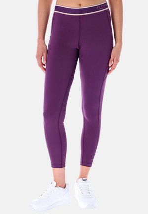 Femme portant des leggings moulants violets avec une taille "Ted Baker Sport" et des baskets blanches Reebok, debout devant un fond uni.