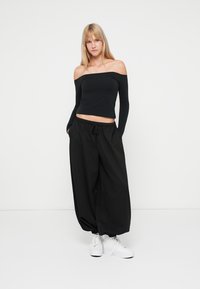 Top nero a spalle scoperte e pantaloni neri larghi con elastico in vita. Modello indossa sneakers bianche. Design pulito e semplice.