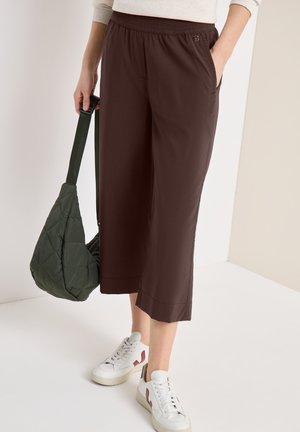 Personne portant un pantalon court marron et des baskets blanches, tenant un sac à bandoulière matelassé vert, debout devant un fond clair.