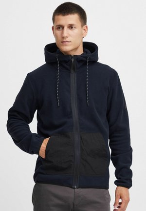 Sudadera con cremallera de color navy que presenta un bolsillo frontal de tela negra texturizada, cordones ajustables en la capucha y un cuerpo de felpa suave.