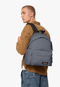 Sac à dos gris avec une texture lisse, sangles noires réglables, poche avant et patch de logo. Porté sur une épaule par une personne.