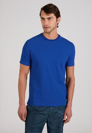 Homme aux cheveux bruns courts portant un t-shirt uni bleu roi à manches courtes et un pantalon sombre à motifs, debout devant un fond uni.