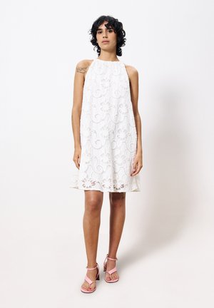 Weißes Spitzenkleid mit hohem Ausschnitt, aufwändigen wirbelnden Mustern und ausgestellter Silhouette. Kombiniert mit rosa Absatzsandalen.
