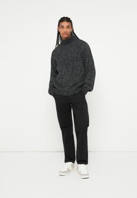 Sort og gråspraglet strikket turtleneck sweater parret med sorte cargo-bukser og hvide sneakers med beige detaljer. En simpel, afslappet påklædning.