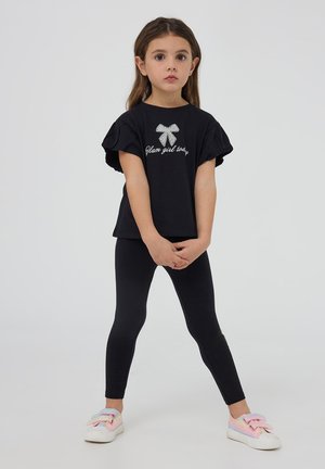 T-shirt nera con maniche arricciate e scritta in strass "Glam girl today", abbinata a leggings neri e sneakers colorate con fiocchi.