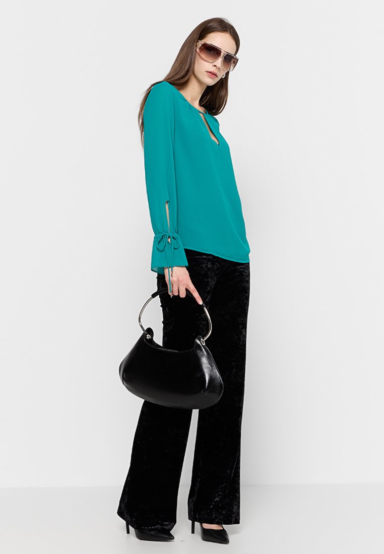 Türkise Bluse mit Bindebändern an den Ärmeln, kombiniert mit schwarzen Samt-Variation-Hosen. Das Model hält eine schwarze Hobo-Tasche und trägt übergroße Sonnenbrillen.