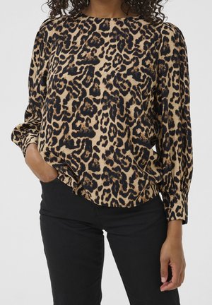 Femme portant un chemisier à manches longues beige et noir imprimé léopard, associé à un pantalon noir, une main dans la poche et des cheveux bouclés visibles.