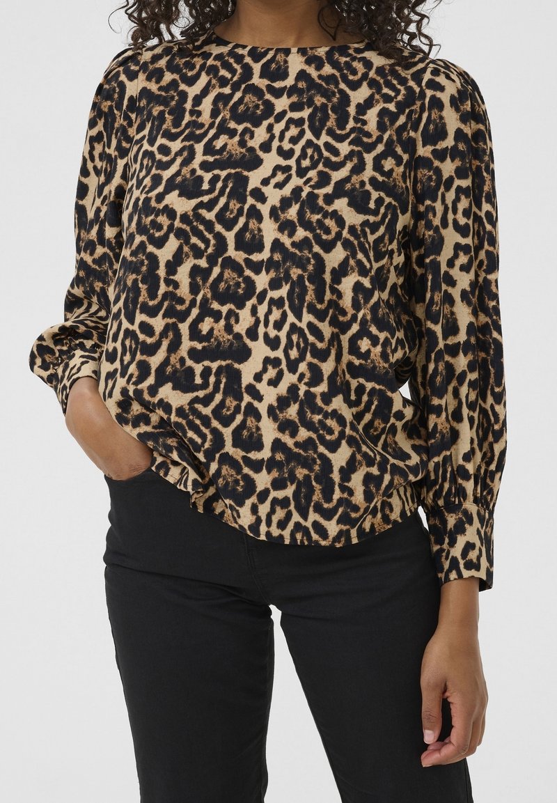 Femme portant un chemisier à manches longues beige et noir imprimé léopard, associé à un pantalon noir, une main dans la poche et des cheveux bouclés visibles.
