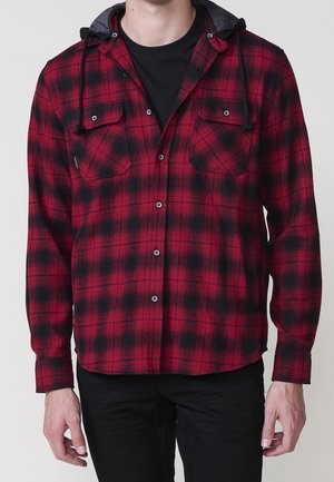 Homme portant une chemise en flanelle à carreaux rouge et noir avec des cordons de capuche noirs par-dessus un t-shirt noir et un pantalon noir.