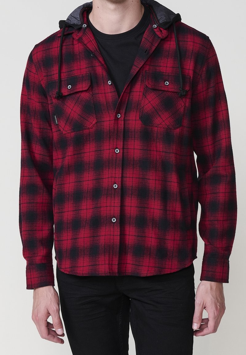 Homme portant une chemise en flanelle à carreaux rouge et noir avec des cordons de capuche noirs par-dessus un t-shirt noir et un pantalon noir.