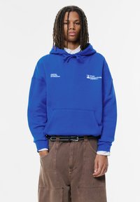 Felpa blu con cappuccio e cordini, caratterizzata da testo bianco e dettagli con logo. Abbinata a pantaloni marroni larghi e una cintura nera con borchie.