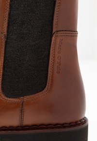 Botte chevillère en cuir marron avec un panneau en tissu noir texturé, des coutures détaillées et un logo de la marque sur le côté. Semelle extérieure en caoutchouc visible.