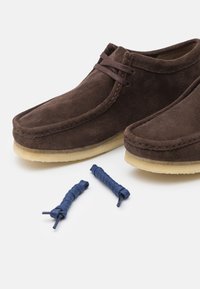 Chaussures en daim marron avec une forme douce et arrondie et des semelles en caoutchouc. Deux lacets bleus au fini texturé sont placés à côté des chaussures.