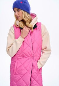 Prošívaná růžová vesta s černými doplňky přes krémovou mikinu; má velké knoflíky, kapsy a diamantový vzor textury. Levandulová čepice.