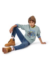 Sweatshirt azul claro com design gráfico, detalhes azuis e verdes, jeans de ganga, botas de tornozelo castanhas e um gorro de malha castanho.