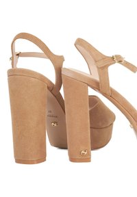 Nude suede high-heeled sandals with a platform, thick block heels, ankle strap, and gold hardware accents. Smooth texture and rounded shape.