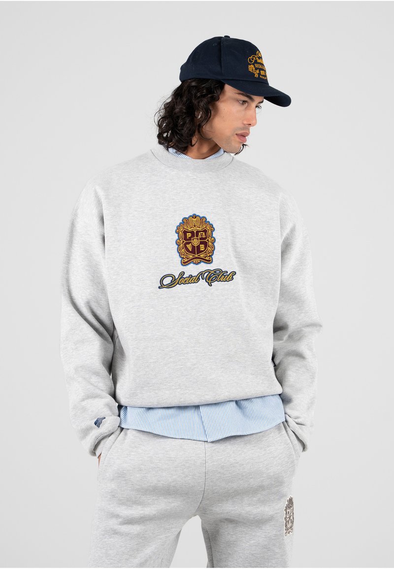 Grijze sweatshirt met een ronde halslijn en geborduurd logo. Gecombineerd met een lichtblauw gestreept shirt eronder en grijze joggingsbroek. Marineblauwe pet.