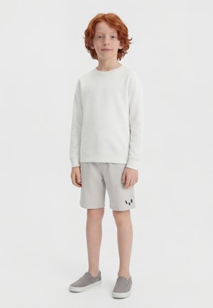 Boy with red hair wearing a white long-sleeve sweatshirt, light gray shorts with a logo, and gray slip-on shoes standing against a plain background.