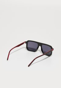 Ochelari de soare negri, cu formă octogonală, lentile închise la culoare și brate subțiri tăiate roșu. Prezintă un design geometric și un finisaj lucios.