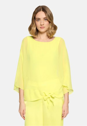 Kvinde med skulderlangt, bølget hår iført en løs, gennemsigtig gul bluse med vide ærmer og en bundet sløjfe i taljen.