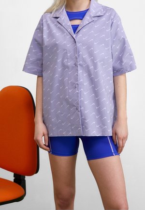 Personne portant une chemise lavande à boutons avec des motifs blancs en forme de virgule sur un short de sport bleu, debout à côté d'une chaise de bureau orange.