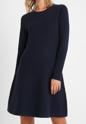 Femme portant une robe bleu marine foncé à manches longues, longueur genoux, coupe évasée, sur un fond blanc uni.