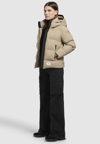 Beige Steppjacke mit Kapuze und schwarzen Akzenten, mit Seitentaschen und einem Logo-Patch. Trägt schwarze Cargo-Hosen und Schuhe.