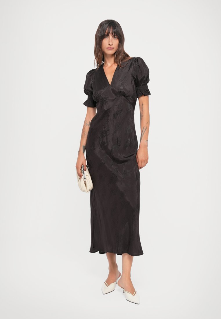 Robe maxi noire en satin avec des manches courtes à volants, décolleté en V profond, taille cintrée et silhouette fluide. Associée à un sac à main blanc texturé.