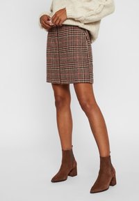 Vrouw in een beige gebreide trui, een geruite knie-lange rok en bruin suède enkellaarsjes staat tegen een effen achtergrond.