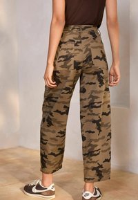 Camo cargo broek in olijf- en zwarttinten, met een ontspannen pasvorm, hoge taille en opgestikte zakken. Gecombineerd met sportieve bruine en witte schoenen.