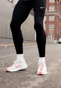 Zwarte sportleggings gecombineerd met lichtgrijze Nike sneakers met roze accenten en witte sokken, geplaatst op een gestructureerd asfaltoppervlak.