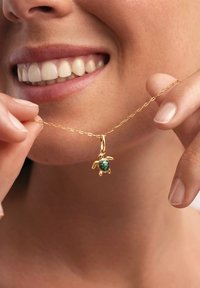 Collar de cadena de oro con un colgante de tortuga que tiene un caparazón texturizado en verde, sostenido por una mano cerca de la boca, sobre un fondo neutro.