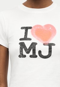 Λευκό μπλουζάκι με μαύρο κείμενο "I MJ" και μεγάλο ροζ καρδιά με υφή μεταξύ των "I" και "MJ", που φοράει ένα άτομο με σκούρο δέρμα.