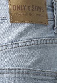 Jasně modrá denimová látka s viditelným švem a hnědou koženou nášivkou s nápisem "ONLY & SONS" a "PASSIONATE JEANS MAKERS."