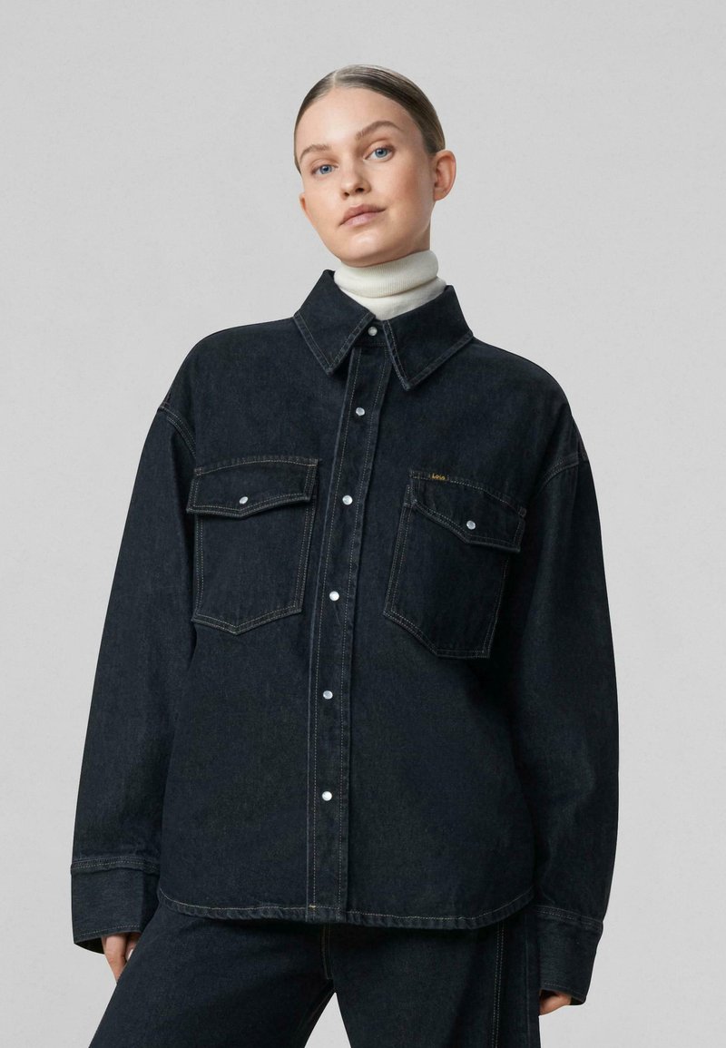 Overshirt in denim scuro con un ampio colletto, due tasche sul petto e chiusure con bottoni bianchi. Presenta una vestibilità comoda e dettagli di cucitura in evidenza.