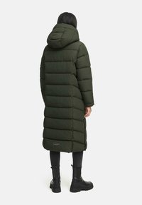 Lange, donkergroene donsjack met capuchon en gewatteerd design. Heeft een logo aan de zoom en wordt gecombineerd met zwarte enkellaarzen en jeans.