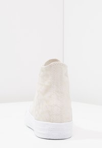 Beżowy wysoki sneaker z teksturowanym materiałem, białą gumową podeszwą i wytłoczonym logo "CONVERSE ALL STAR" na pięcie. Gładki, minimalistyczny design.