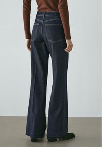 Jeansy o ciemnym odcieniu z wysokim stanem i rozszerzanymi nogawkami, z widocznym przeszyciem, dwoma tylnymi kieszeniami oraz gładkim wykończeniem, noszone z dopasowanym brązowym topem.