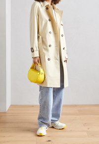 Beige värinen trenssitakki kaksirivisellä napituksella, isoilla napeilla ja vyötärönauhalla. Vaaleansiniset väljät farkut ja valkoiset tennarit keltaisilla yksityiskohdilla.