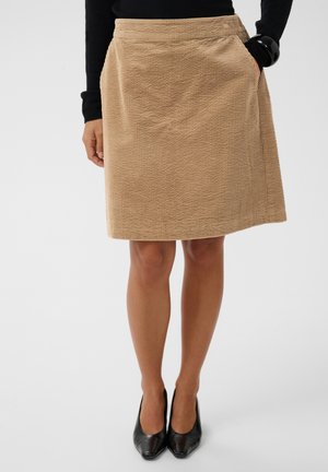 Beige corduroy-rok tot op de knieën gedragen met een zwarte top en zwarte dichte hakken, handen rustend langs de zij.