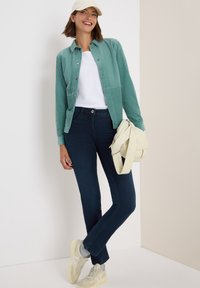 Glunderende vrouw in een crèmekleurige pet, met een teal ribfluwelen jack over een wit shirt, donkere jeans, beige sneakers en een crèmekleurige handtas.