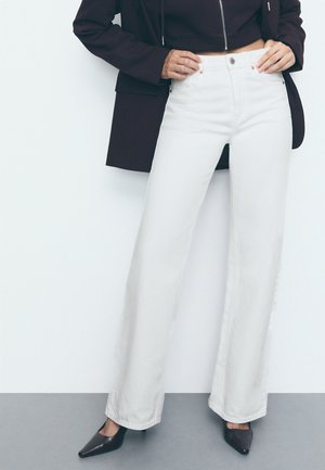 Personne portant un jean blanc taille haute à jambes larges avec bouton et fermeture éclair, blazer noir et talons pointus noirs sur un sol gris.