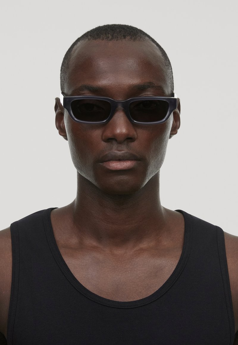 Gafas de sol negras rectangulares con montura delgada, llevadas por una persona con una camiseta sin mangas negra. Textura suave y diseño minimalista.