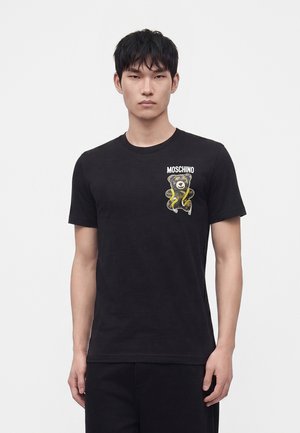 Άνδρας που φοράει μαύρο t-shirt Moschino με εικόνα αρκουδάκι και κίτρινο φίδι στο αριστερό στήθος, στέκεται μπροστά σε απλό φωτεινό φόντο.