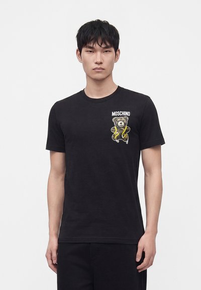 Άνδρας που φοράει μαύρο t-shirt Moschino με εικόνα αρκουδάκι και κίτρινο φίδι στο αριστερό στήθος, στέκεται μπροστά σε απλό φωτεινό φόντο.